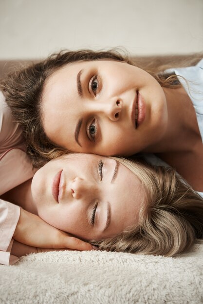 Mother and daughter looking like best friends. Vertical shot of young attractive woman lying on head of another girl,  and smiling with relaxed and calm expression, feeling happy