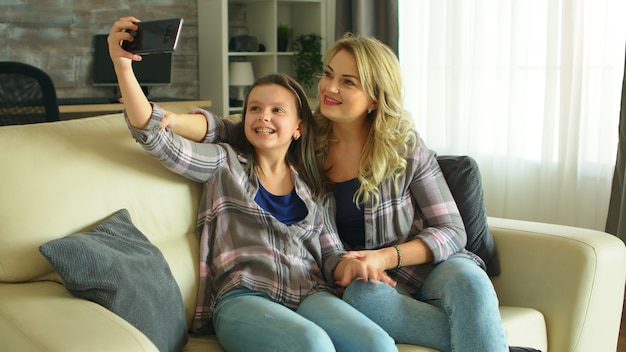 Mother and daughter making funny faces while taking a selfie sitting on the couch in living room.