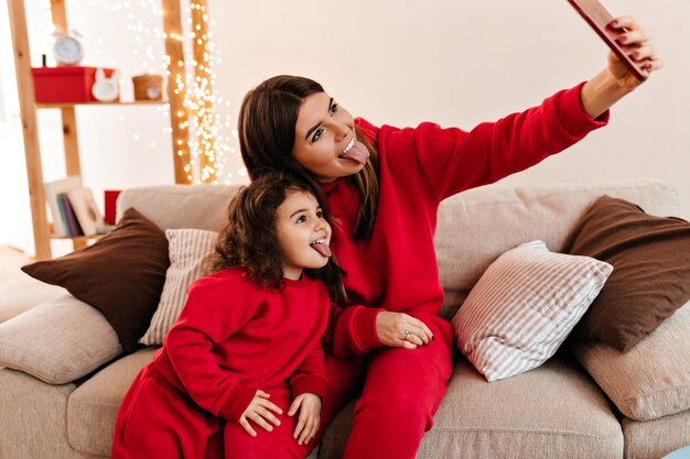 Mother and daughter posing with tongues out. Stylish mom taking selfie with kid at home.