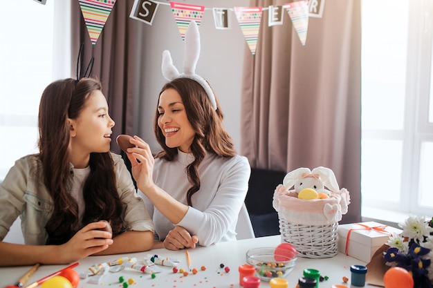 Mother and daughter prepare for easter. they sit at table in room. young woman feed girl with chocolate egg. kid keep mouth opened. decoration and paint with sweets on table.