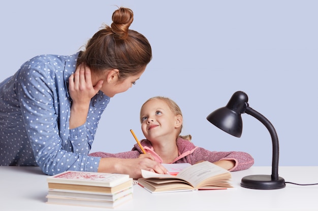 mother and daughter sitting at table surrounded by books looking at each other with love, doing homework together, mummy helps little girl to do sums. Children, school, education concept.