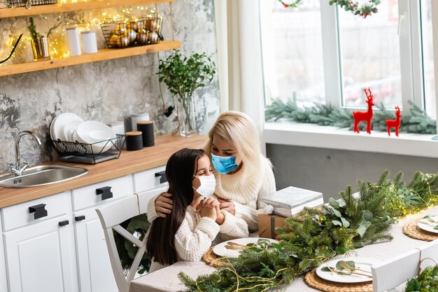 어머니와 딸은 집에서 축하하는 동안 크리스마스 트리 아래에서 의료용 마스크를 쓰고 있습니다.