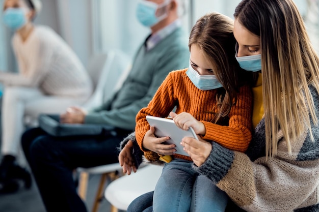 Mother and daughter with face masks using touchpad while waiting at medical clinic