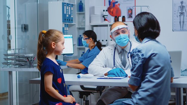 Mother explaining to doctor girl symptoms during coronavirus in medical office. Pediatrician specialist in medicine with mask providing health care services consultation, treatment in hospital cabinet