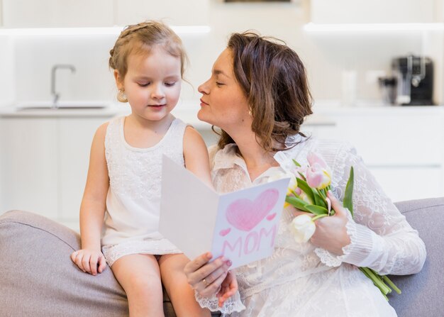 Mother giving kiss to her cute little daughter holding greeting card and flower bouquet