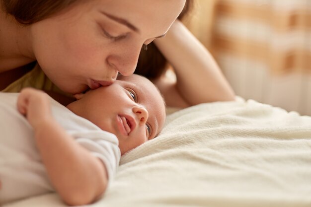 Mother and her newborn baby together lying on bed on blanket. Happy mother kissing and hugging baby, kid looking away and studying outward things. Maternity and parenthood.