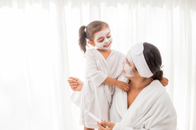 Mother and her young pretty daughter having fun in the bedroom and making clay facial masks