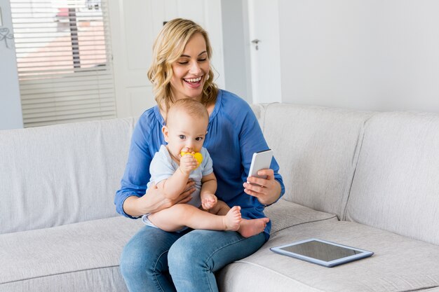 Mother holding baby boy and using mobile phone in living room