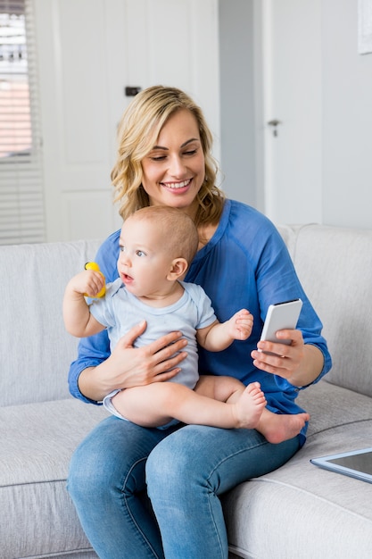 Mother holding baby boy and using mobile phone in living room
