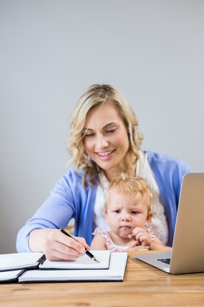 Mother holding baby girl while writing notes in personal organizer