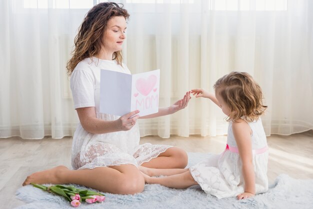 Mother holding greeting card sitting near her daughter on carpet at home