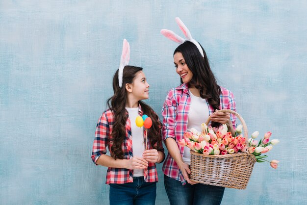 Mother holding tulips basket looking at girl holding easter eggs in hand against blue background