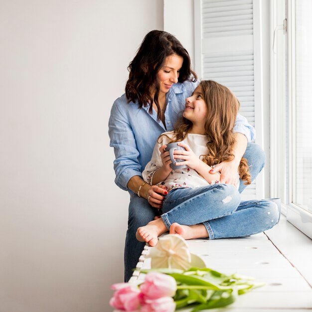 Mother hugging daughter from behind on window sill