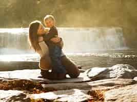 Free photo mother hugging her son in a park surrounded by greenery and a waterfall under the sunlight