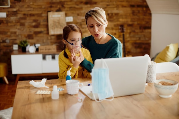 Free photo mother inhaling her small daughter with nebulizer while having video call with a doctor from home