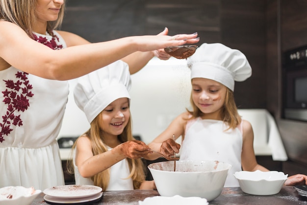 Mother sifting cocoa powder with strainer in bowl while happy girls preparing food