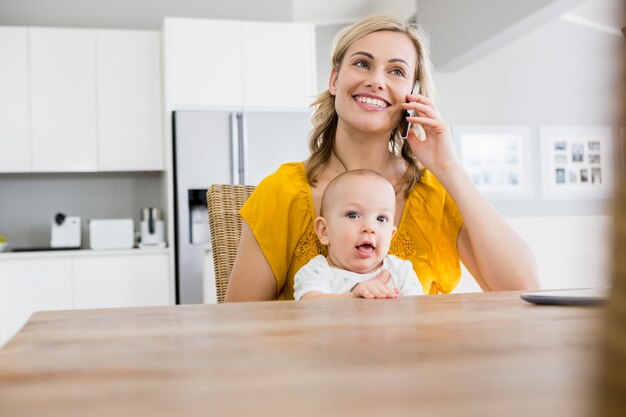 Mother talking on mobile phone with baby boy in kitchen