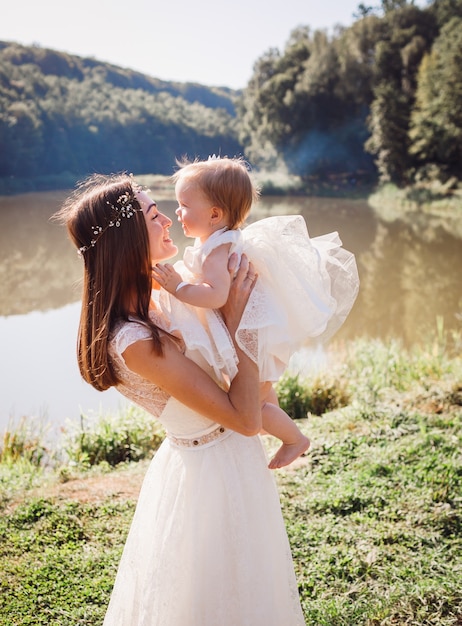 Mother in white dress plays with her charming daughter in white gawn before a lake