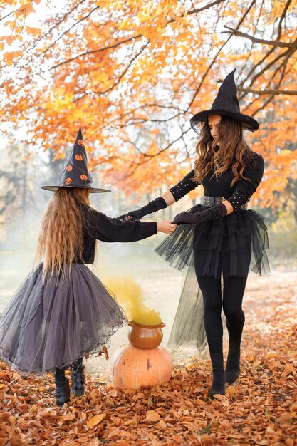 Мать-ведьма и ее дочь варят зелье в осеннем лесу. Мать учит дочь. Молодая женщина и маленькая девочка в костюмах Хэллоуина ведьм и конусных шляпах.