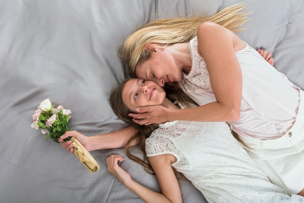 Мать с цветами обнимает дочь на кровати