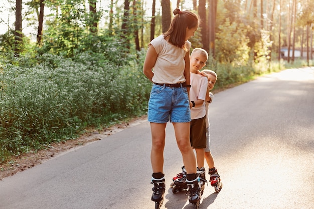 Mother with sons spending time together in summer park, family rollerblading, active pastime, children with mother roller skating outdoor on asphalt road.