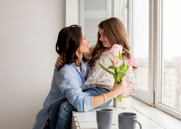 Мать с тюльпанами обнимает дочь у окна