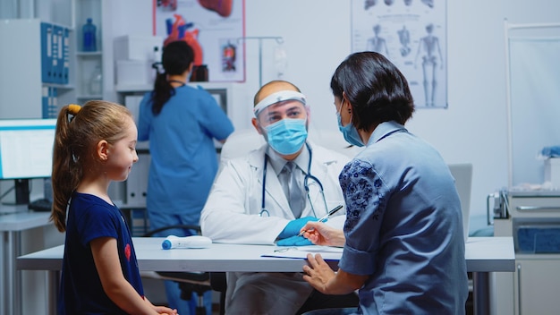 Mother writing child treatment on clipboard sitting in medical office. Pediatrician specialist in medicine with mask providing health care services, consultation, treatment in hospital during covid-19