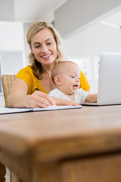 Mother writing on a diary while baby boy playing with laptop in kitchen