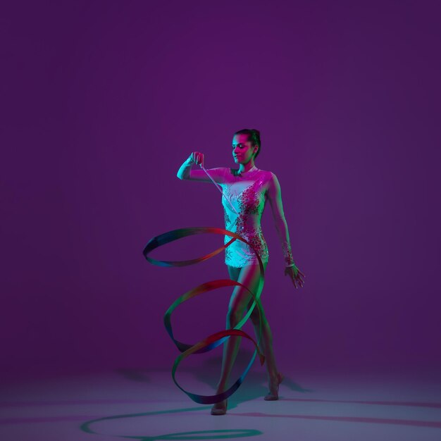 モーション。若い女性アスリート、新体操アーティストのダンス、ネオンの光の中で紫色のスタジオの背景に分離されたテープでトレーニング。機器で練習している美少女。パフォーマンスの優雅さ。