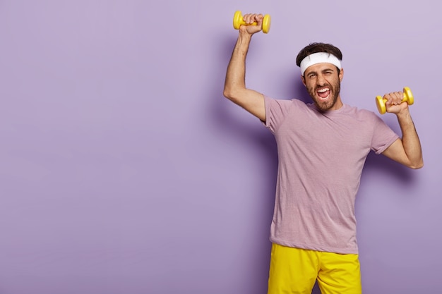 Motivated sportsman trains muscles, raises yellow dumbells, wears headband, casual outfit, being active