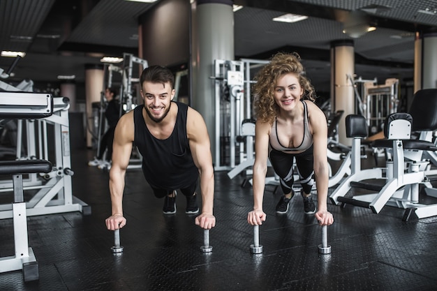 Motivated young blond woman and man trainer in middle of workout, standing in plank with dumbbells.