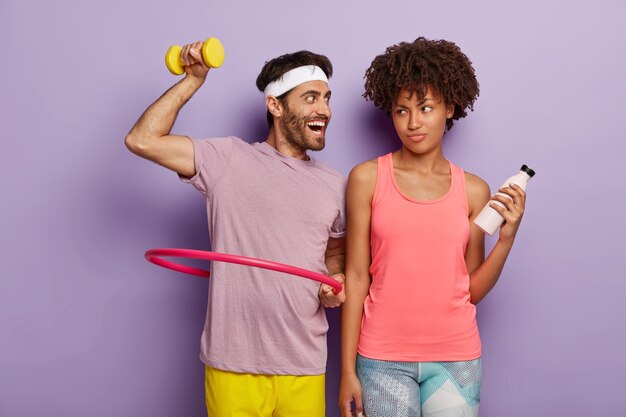 Motivated young man exercises with hula hoop, raises dumbbell, has glad expression, wears white headband and t shirt and displeased woman stands with bottle of water, has fitness training