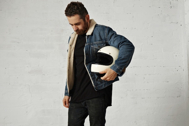 motor biker wears shearling denim jacket and black blank henley shirt, holds vintage beige motorcycle helmet, looking down, isolated in center of white brick wall