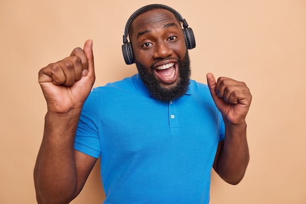 Move your body. Glad bearded man dances in wireless headphones enjoys favorite song sings happily dressed casually isolated over beige studio wall uses best music app