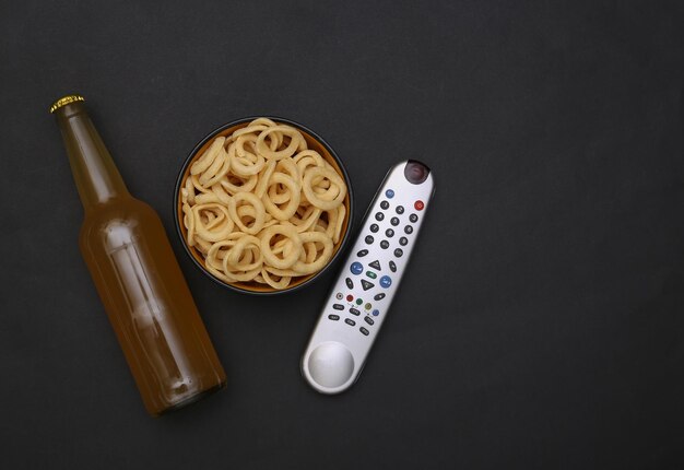 Movie time. bottle of beer and bowl with corn rings snack, tv remote on black background. top view