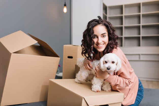 小さな犬と一緒に若いきれいな女性の新しいアパートに移動します。ベッドの上で身も凍るように、ペットと一緒にカートンボックスを囲み、笑顔で、積極性を表現する