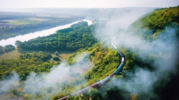 Moving train on railway with high column of smoke, flowing river, hills and railway on the foreground