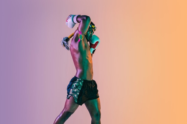 Free photo muay thai. young man exercising thai boxing on gradient background in neon light. fighter practicing, training in martial arts in action, motion. healthy lifestyle, sport, asian culture concept.