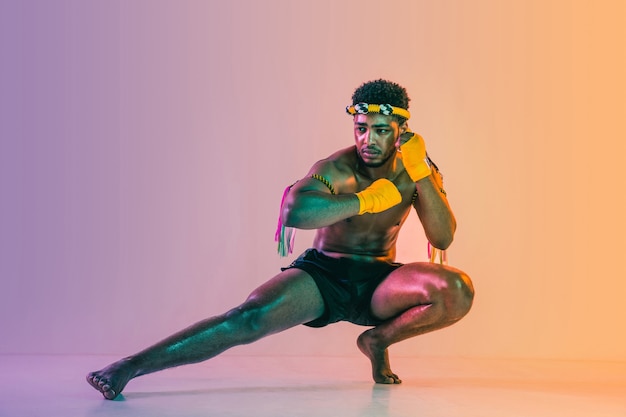 Muay thai. Young man exercising thai boxing on gradient background in neon light.