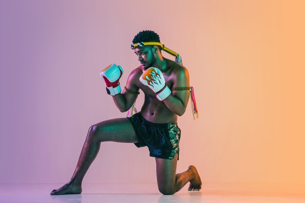 Muay thai. Young man exercising thai boxing on gradient wall in neon light. Fighter practicing, training in martial arts in action, motion. Healthy lifestyle, sport, asian culture concept.