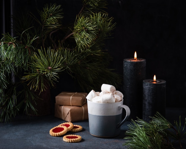 Mug with cocoa, marshmallow and cinnamon on a dark blue background with fruit cookies, lit candles, gifts and fir tree. dark and mood image