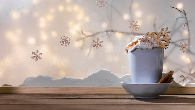 Mug with toy snowflake on plate with cookies on wood table near bank of snow and fairy lights
