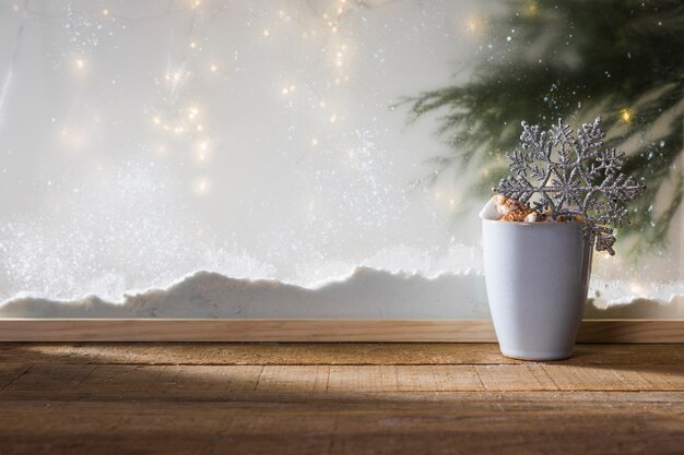 Mug with toy snowflake on wood table near bank of snow, fairy lights and fir twig