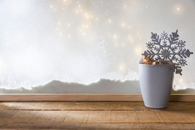 Mug with toy snowflake on wood table near bank of snow and fairy lights