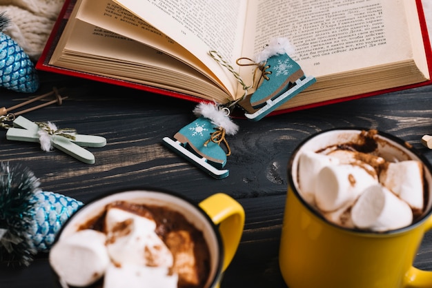 Mugs with marshmallows and drinks near Christmas decorations and book