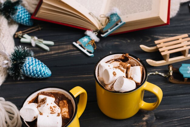 Mugs with marshmallows near Christmas decorations and book 