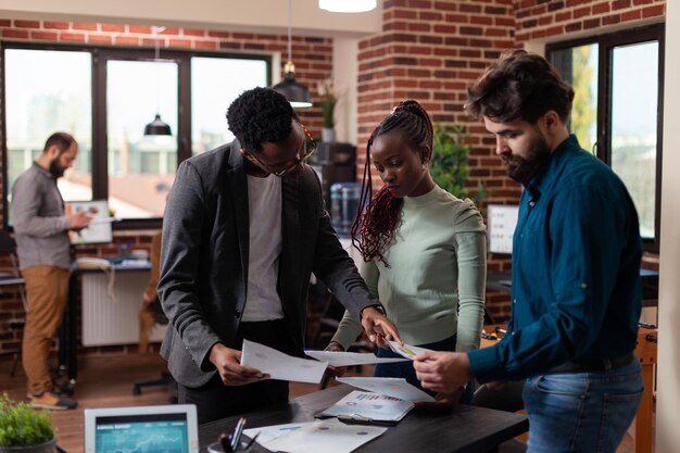 Multi-ethnic businesspeople discussing company collaboration analyzing papers with statistics charts working in brick wall office. Diverse team brainstorming ideas for business strategy
