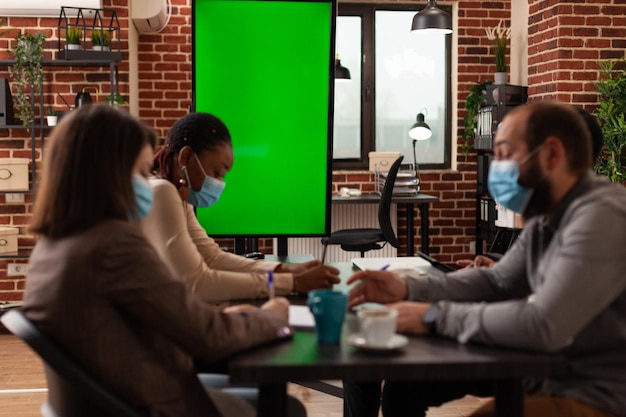スタートアップ企業の会議室でのビジネスプレゼンテーションで働くコロナウイルスに対する医療用フェイスマスクを持つ多民族のビジネスマン。分離されたディスプレイを備えたグリーンスクリーンクロマキーモニターのモックアップ