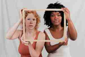 Free photo multi ethnic female friend looking at camera through wooden frame against grey backdrop