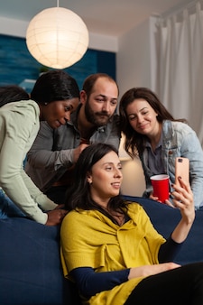 自宅​の​居間​で​の​ビデオ​通話中​に​スマート​フォン​で​結合して​話している​多​民族​の​友人​。​居間​で​夜​遅く​ソファ​に​座って​一緒​に​時間​を​過ごす​多​民族​の​人々​の​グループ​。